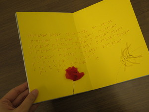 イタリアのさわる絵本。紙製で縦長の判型。黄色い紙に点字が打たれており、左下には赤い花が貼られている。